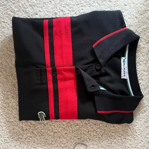 Svart och röd pikétröja Lacoste L - Svart pikétröja från Lacoste i storlek L med två breda röda ränder framtill och röda detaljer på kragen. Klassisk pikékrage med knappar och den ikoniska krokodilloggan broderad på bröstet. Tröjan är tillverkad i bomull och har korta ärmar.