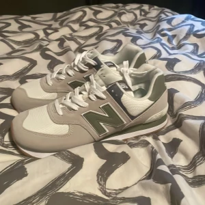 New Balance 574 sneakers grå/vit - Snygga New Balance 574 sneakers i grått och vitt med gröna detaljer. Skorna har en klassisk rund tå, vit snörning och en slitstark gummisula med grovt mönster. Ovandelen är i mesh och mocka för både stil och komfort. Perfekt för dig som gillar retrovibe och streetstyle.