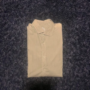 Beige skjorta från Stacius - Säljer en stilren beige skjorta från Stacius Heritage Limited Edition. Skjortan har klassisk krage och knäppning framtill med vita knappar. Perfekt för dig som gillar enkel och clean stil. Materialet känns lätt och skönt mot huden.