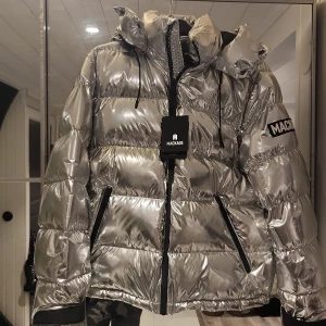 Silver pufferjacka från Mackage - Säljer en riktigt fet silverfärgad pufferjacka från Mackage. Jackan har en glansig finish, huva, svarta dragkedjor och tydlig logga på ärmen. Perfekt för dig som vill sticka ut i vinterkylan. Fodrad och med snygga detaljer som dragsko vid huvan.