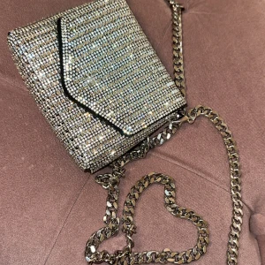 Glittrig kuvertväska med kedja - Supersnygg kuvertväska täckt av glittrande stenar och med en silvrig kedja som axelrem. Väskan har en svart kant runt locket och är perfekt för att lyfta vilken outfit som helst. Liten och smidig storlek, perfekt för mobilen och det viktigaste.