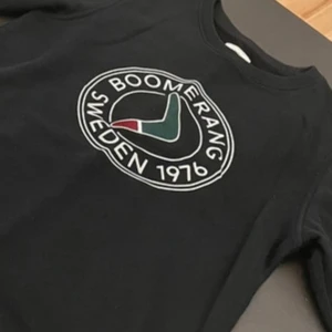 Svart sweatshirt från Boomerang - Svart sweatshirt från Boomerang med stort tryck på bröstet där det står 'Boomerang Sweden 1976' och en boomerang-symbol i grönt, vitt och rött. Tröjan har rund hals och långa ärmar, perfekt för en chill och stilren look.