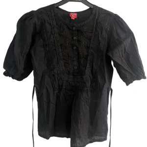 Svart blus med spets och puffärm - Svart blus från Koola anna med puffiga korta ärmar, spetsdetaljer och knappar framtill. Blusen har rund halsringning och knytband i midjan. Tillverkad i tunn bomull med diskret struktur i tyget.