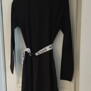 Svart långärmad klänning med bälte - Svart långärmad klänning från Missguided i enkel och stilren design. Klänningen har ett vitt bälte med MSGD-tryck som ger en cool kontrast och markerar midjan. Perfekt för dig som gillar minimalistisk stil och vill ha något bekvämt men ändå trendigt.