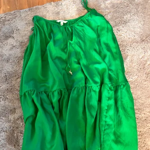 Grön sommarklänning från H&M - Säljer en grön klänning från H&M i storlek M. Klänningen har tunna axelband, volangdetaljer och knytning framtill. Den är luftig och har en glansig finish som ger en fräsch vibe. Perfekt för varma dagar när du vill sticka ut lite extra.