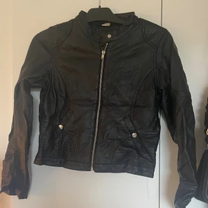 Svart skinnjacka med dragkedja - Säljer en svart skinnjacka med coola sömdetaljer och silvriga knappar vid fickorna. Jackan har en rak passform, lång ärm och stängs med dragkedja framtill. Perfekt för dig som vill ha en edgy look till vardags. Fråga gärna om du vill veta mer om storlek!