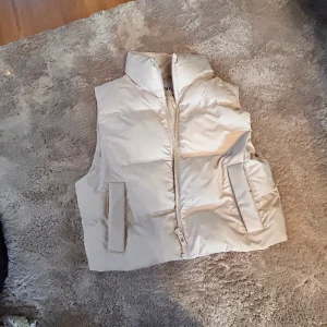 Beige dunväst från Zara XS - Snygg beige dunväst från Zara i storlek XS. Västen har hög krage, två fickor framtill och stängs med dragkedja. Perfekt att slänga över en hoodie eller tröja för en trendig look.