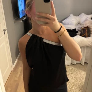 Svart oneshoulder topp med silverdetalj - Snygg svart oneshoulder topp med glittrigt silverband längs axeln och halsringningen. Toppen har en lös passform och är tillverkad i ett mjukt, lätt material som ger ett elegant fall. Perfekt för dig som vill sticka ut med en cool och modern look.