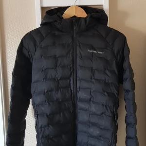 Svart dunjacka Peak Performance - Säljer en svart dunjacka från Peak Performance med huva och dragkedja framtill. Jackan har quiltad design och två sidofickor med dragkedja. Perfekt för kyliga dagar och enkel att matcha med allt.