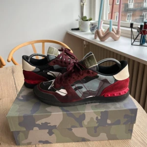 Valentino Garavani Rockrunner sneakers - Snygga Valentino Garavani Rockrunner sneakers med camouflagemönster i vinrött, grått, svart och vitt. Skorna har detaljer i mocka och skinn, samt ikoniska nitar på den röda hälen. Platt sula och snörning framtill. Perfekt för dig som gillar statement-sneakers. Skriv vid funderingar!