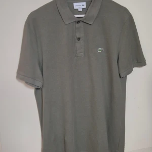 Olivgrön Lacoste piké L - Snygg olivgrön pikétröja från Lacoste använd endast 3-4 gånger. Klassisk krage med tre knappar och den ikoniska krokodilloggan broderad på bröstet. Tillverkad i mjuk bomull som andas och har korta ärmar. Perfekt för dig som gillar stilren och enkel design.