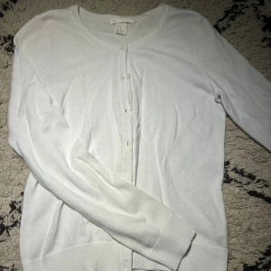 Vit kofta med knappar från H&M - En stilren vit kofta från H&M Basic med rund hals och långa ärmar. Koftan har knappar framtill. Perfekt att slänga över en t-shirt eller linne för en clean look.