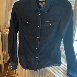 Mörkblå skjorta från G-Star RAW XS - Snygg mörkblå skjorta från G-Star RAW i klassisk jeansstil. Skjortan har långa ärmar, vita tryckknappar och två bröstfickor med lock. Materialet är kraftigt och ger en cool denimlook. Perfekt för dig som gillar streetwear och stilrena detaljer.