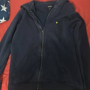 Marinblå hoodie från Lyle & Scott - Snygg marinblå hoodie från Lyle & Scott med gul broderad logga på bröstet. Tröjan har huva, dragkedja framtill och två stora fickor. Perfekt för chill dagar och enkel att matcha med jeans eller joggers. Storlek 14-15