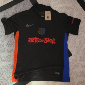 FC Barcelona x Nike Lamine Yamal tröja - Snygg FC Barcelona x Nike fotbollströja med Lamine Yamal och nummer 10 på ryggen. Svart bas med röda och blå detaljer, samt orange tryck på sidorna. Dri-FIT material som andas, korta ärmar och coolt Cactus Jack-tryck på bröstet. Perfekt för dig som älskar Barca och streetstyle.