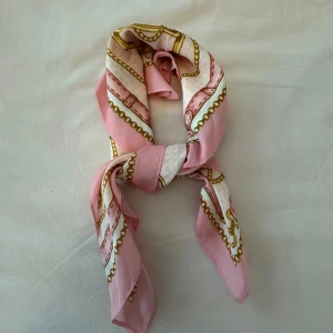 Rosa och vit scarf med kedjemönster - Snygg fyrkantig scarf från Vero Moda i rosa och vitt med detaljerat kedje- och remmönster i guld. Tillverkad i mjuk polyester som känns lyxig mot huden. Perfekt att knyta runt halsen eller håret för en chic look.