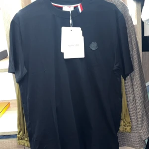 Moncler tshirt - Svart t-shirt från Moncler med diskret logopatch på bröstet och klassisk rund hals. Kragen har snygga detaljer i rött, vitt och blått. T-shirten är tillverkad i mjuk bomull och har korta ärmar, perfekt för en clean och stilren look.