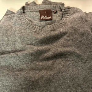 Grå stickad tröja från Oscar Jacobson - Mysig grå stickad tröja från Oscar Jacobson i klassisk modell. Tröjan har rund halsringning med ribbade detaljer och är perfekt för kyliga dagar. Enkel och stilren design som funkar till det mesta. S