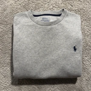 Grå sweatshirt från Polo Ralph Lauren - Snygg grå sweatshirt från Polo Ralph Lauren med klassisk rund hals och mörkblå broderad logga på bröstet. Tröjan har långa ärmar, ribbade muddar och är tillverkad i mjuk bomullsmix som är perfekt för chill dagar.