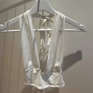 Vit omlott halterneck topp - Säljer en vit omlott topp med djup v-ringning och knyt i nacken. Toppen är ärmlös och croppad, perfekt för varma dagar. Tillverkad i mjukt och lätt material som ger en snygg siluett.