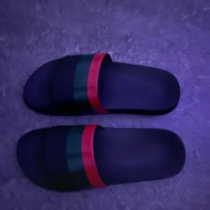 Svarta sandaler med röd och grön rand - Svarta sandaler med bred rem i klassisk röd och grön rand. Snygg och enkel design med platt sula och öppen tå. Perfekta för sommaren och chill dagar. Tillverkade i syntetmaterial för en lätt känsla på foten.