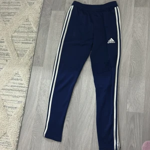 Marinblå Adidas träningsbyxor XS - Snygga marinblå träningsbyxor från Adidas med vita ränder längs benen och logga på låret. Byxorna har dragkedja vid bensluten och är gjorda i mjuk polyester som är perfekt för sport och aktivitet. Passformen är slim och midjan är normal.