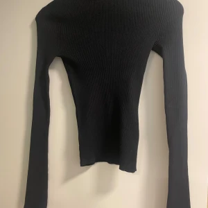 Svart ribbad långärmad topp från EcoVero - Säljer en svart, ribbad långärmad topp från EcoVero med rund, öppen rygg. Toppen är tight i passformen och har ett stilrent, minimalistiskt uttryck. Materialet är mjukt och stretchigt syntet, perfekt för dig som gillar en clean look med en twist.