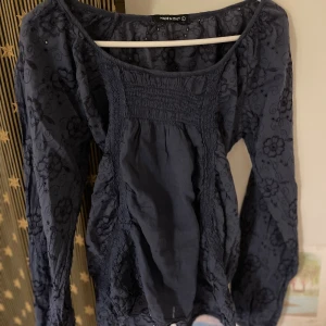 Mörkblå blommig blus med knyt - Supersöt mörkblå blus med broderade blommor och smockdetaljer framtill. Blusen har långa ärmar, rundad hals och ett knytband i midjan för extra stilpoäng. Materialet känns lätt och luftigt, perfekt för dig som gillar romantiska vibes.