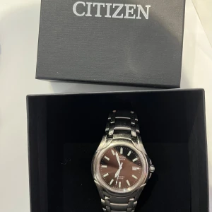 Citizen klocka  - Snygg klocka från Citizen med mörk urtavla och datumvisning. Boetten och länken är i titanium, vilket gör klockan både lätt och slitstark. Den har safirglas, vilket ger extra skydd mot repor. Klockan är i fint begagnat skick med normalt bruksslitage (se bilder). Den fungerar som den ska och håller tiden bra. En riktigt kvalitetsklocka med klassisk design som passar både till vardag och finare tillfällen. ✅ Quartz-urverk ✅ Vattentålig (10 BAR - alltså 100 meter) ✅ Datumvisning ✅ Citizen ask