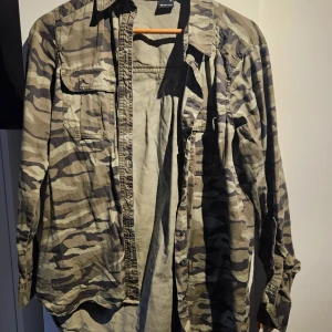 Camouflage skjorta från Gina Tricot - Snygg camouflage-mönstrad skjorta från Gina Tricot i storlek 36. Skjortan har två bröstfickor, knappar framtill och långa ärmar med knappdetalj. Perfekt för dig som gillar streetstyle och vill sticka ut lite extra.