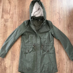 Olivgrön parkas från H&M Divided / Olive green parka from H&M Divided - Olivgrön parkas med huva från H&M Divided. Jackan har dragsko i midjan, två stora fickor med lock och knappar samt mysigt foder i huvan. Yttermaterial i bomull och foder i polyester. Perfekt för kyliga dagar och snygg till jeans eller leggings.