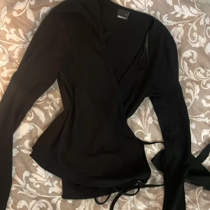 Svart omlottopp från Gina Tricot.  - Svart omlottopp från Gina Tricot i storlek S. Topp med långa ärmar och knytband i midjan som ger en snygg siluett. Stilren och enkel design, perfekt att matcha med jeans eller kjol. Materialet känns mjukt och följsamt mot kroppen. Använd en gång och är i nyskick.🌸