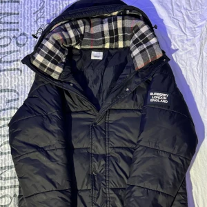 Svart pufferjacka från Burberry - Svart pufferjacka från Burberry med avtagbara ärmar och klassiskt rutigt foder i huvan. Jackan har dragkedja och tryckknappar framtill samt två fickor med dragkedja. På vänster ärm finns Burberry London England-tryck. Perfekt för kalla dagar. 🆘Jag behöver pengar därför jag vill sälja så fort 🆘
