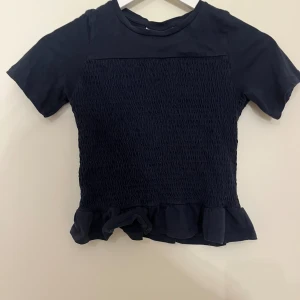 Mörkblå topp från Gina Tricot - Mörkblå topp från Gina Tricot med korta ärmar och smockad front. Toppen har en söt volangkant nedtill som ger en extra detalj. Tillverkad i mjukt bomullsmaterial och har en enkel, stilren look som passar till mycket. Har liten defekt (se bild) men syns ej när man har på sig eftersom håret brukar täcka. Storlek 134/140 men passar mig som kan ha xxs och xs.