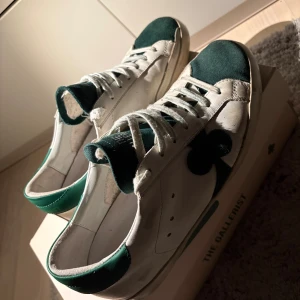 The Gallerist x AMI vita och gröna sneakers - Snygga sneakers från AMI x The Gallerist i vitt skinn med gröna mockadetaljer på tå, häl och plös. Klassisk låg modell med snörning och svart platt sula. Ikonisk klöverlogga på sidan och text på plösen. Perfekt för dig som gillar unik design.