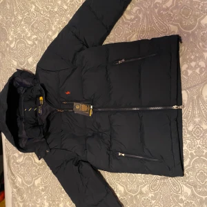 Mörkblå pufferjacka från Polo Ralph Lauren - Säljer en mörkblå pufferjacka från Polo Ralph Lauren med huva och dragkedja. Jackan har orange broderad logga på bröstet och två fickor med dragkedja. Perfekt för kalla dagar och riktigt snygg streetstil.