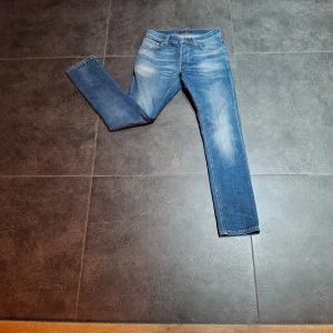 Blå jeans från Nudie Jeans, W27 L30 - Snygga blå jeans från Nudie Jeans med klassisk femficksdesign och orangea kontrastsömmar. Modellen har en rak passform och normal midja. Materialet är slitstarkt denim i bomull. Bakfickan har Nudie Jeans signaturbroderi.
