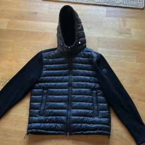 Svart hybridjacka från Colmar, strl 52 - Snygg svart hybridjacka från Colmar Originals med quiltad framsida och huva i pufferstil. Ärmar och rygg är i mjuk stickad kvalitet för extra komfort. Jackan har inga tecken på användning och är använd ett fåtal gånger.pris är inte hugget i sten