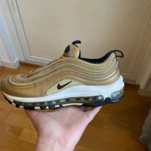 Nike Air Max 97 Gold Metallic 38.5 - Säljer ett par Nike Air Max 97 i färgen Gold Metallic med ikoniska vågformade linjer, synlig Air-sula och röda detaljer på Swoosh-loggan. Skorna har snörning, svart och vit sula samt mesh och syntetmaterial för en sportig vibe. Skickar inom 48 timmar.