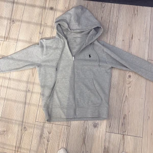 Grå hoodie från Polo Ralph Lauren - Säljer en stilren grå hoodie från Polo Ralph Lauren med dragkedja och huva. Tröjan har det klassiska broderade logotypen på bröstet och är tillverkad i mjuk bomull. Perfekt för chill dagar eller när du vill ha en clean look.