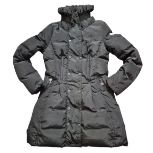 Svart lång dunjacka från Moncler - Lång svart dunjacka från Moncler med hög krage och dubbelknäppning framtill. Jackan har dragkedjor vid fickorna, quiltad design och Moncler-logga på ärmen. Perfekt för kalla vinterdagar och har en snygg, figurnära passform.