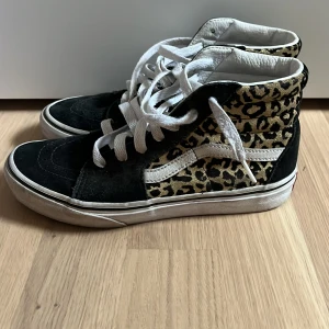 Vans leopardmönstrade sneakers - Säljer ett par Vans high-top sneakers med svart mocka och leopardmönstrade paneler i guld och svart. Skorna har vit sula, vit sidestripe och vita skosnören. 