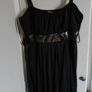 Svart lång linne med dekorerad midja - Svart lång linne från Marina Kaneva i plusstorlek med breda axelband och snyggt dekorerad midja med metalliska detaljer. Klänningen har knytband i ryggen och ett mjukt, fallande tyg som ger en elegant siluett. Perfekt för dig som gillar statement-detaljer.