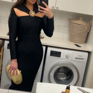 Svart långklänning från Bella Barnett - Supersnygg svart långklänning från Bella Barnett med unik cut-out upptill och en guldig solbrosch med pärla vid halsen. Klänningen har lång ärm, figurnära passform och ett elegant axelparti som ger en cool och modern vibe. Perfekt för dig som vill sticka ut.Normal i storlek använd i några timmar,nyligen köpt