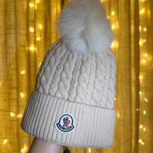 Beige Moncler mössa med päls - Snygg beige kabelstickad Moncler mössa med en stor fluffig pälsboll på toppen. Mössan har bred ribbad uppvikning och den klassiska Moncler-logga framtill. Materialet är av mjukt och tjockt ull, perfekt för kalla dagar. Endast testad på annars helt ny!