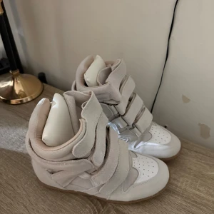 Isabel Marant höga sneakers i beige mocka - Säljer ett par ikoniska höga sneakers från Isabel Marant i ljusbeige mocka med vita detaljer och gummisula. Skorna har flera breda kardborreband, rund tå och plattformskänsla. Perfekt för dig som gillar statement-sneakers med unik design.