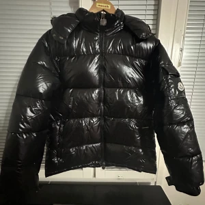 Svart pufferjacka från Moncler - Säljer en svart, glansig pufferjacka från Moncler med klassisk logga på ärmen. Jackan har dragkedja framtill, hög krage och stora vadderade sektioner. Perfekt för kalla dagar och riktigt snygg streetstil.