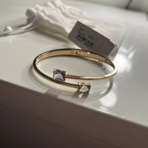 Guldfärgat armband från Lauren Ralph Lauren - Stilrent guldfärgat armband från Lauren Ralph Lauren med två rektangulära, klara stenar i varje ände. Enkel och elegant design som ger en lyxig känsla. Kommer med originalpåse och etikett kvar.