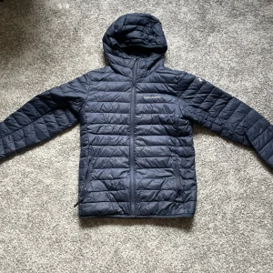 Mörkblå dunjacka Peak Performance S - Säljer en mörkblå dunjacka från Peak Performance i storlek S. Jackan har huva, två dragkedjeförsedda fickor och quiltad design. Perfekt för kyliga dagar och riktigt skön att bära. Klassisk look med diskret logga på bröstet.