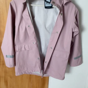 Ljusrosa regnjacka från McKinley - Snygg ljusrosa regnjacka från McKinley med huva och silverfärgade reflexdetaljer på ärmar och rygg. Jackan har tryckknappar framtill, två stora fickor och är tillverkad i syntetmaterial som står emot regn. Perfekt för blöta dagar och har en clean, stilren look.